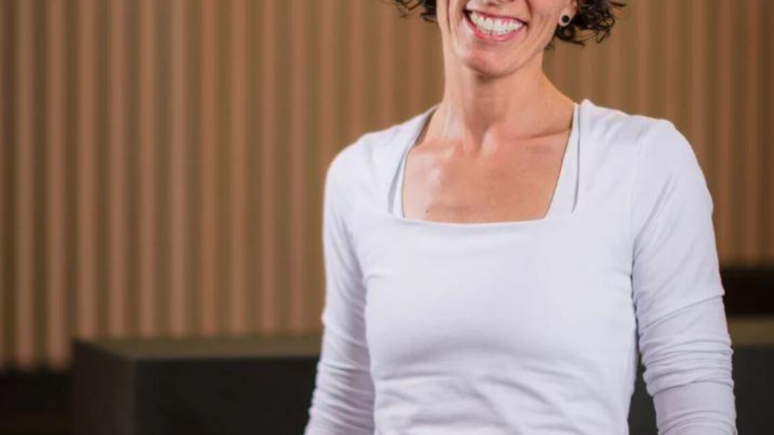 Ärztin mit kurzen lockigen Haaren steht lächelnd vor einem modernen, warm beleuchteten Innenraum; sie trägt ein weißes Langarmshirt und Jeans, im Hintergrund ein unscharfes beleuchtetes Schriftlogo. | © COPRAXIS