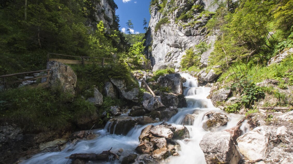 Ein Wasserfall fließt über große Steine in eine grüne Schlucht. Umgeben von hohen Felsen und Bäumen strahlt die Szene natürliche Ruhe aus. | © Gerhard Pilz 