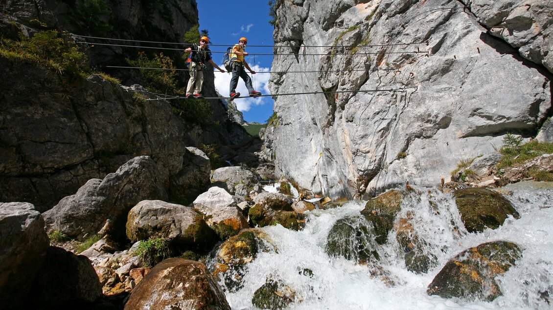 Zwei Personen überqueren eine Hängebrücke über einen sprudelnden Bach in einer felsigen Landschaft. Im Hintergrund sind hohe Felsen und ein klarer blauer Himmel zu sehen. | © photo-austria, Herber Raffalt