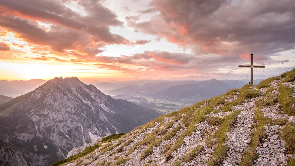 Eine atemberaubende Berglandschaft mit einem Kreuz im Vordergrund. Der Himmel ist von einem warmen Sonnenuntergang erleuchtet. | © Christoph Huber