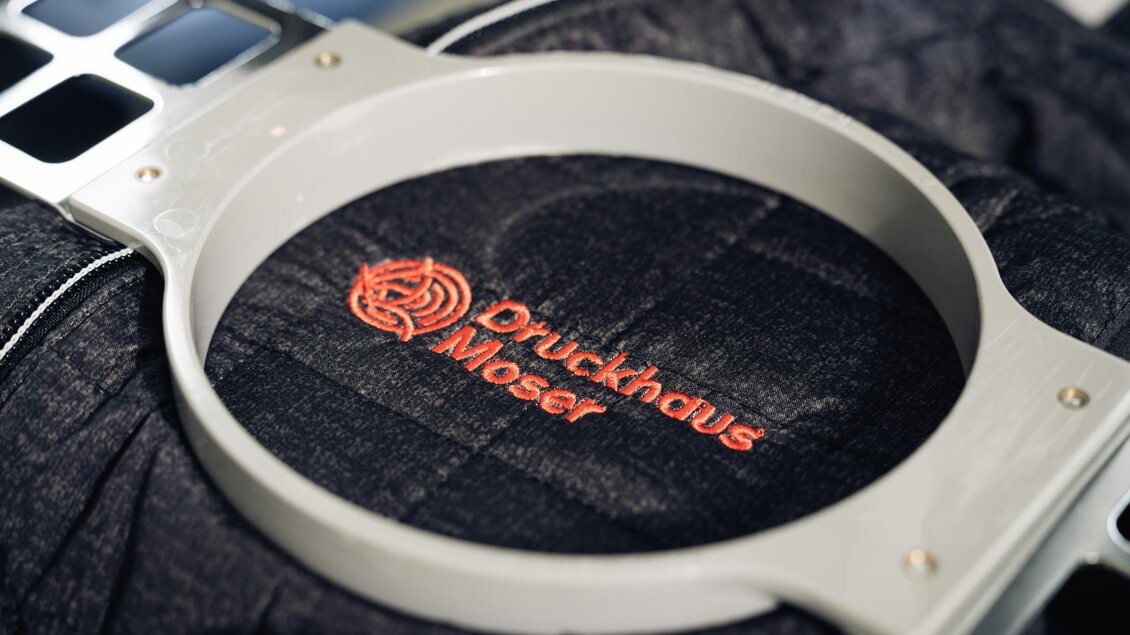 Rote Stickerei mit dem Logo von Druckhaus Moser auf dunklem Textil in einer Stickmaschine. | © Bernd Grosseck