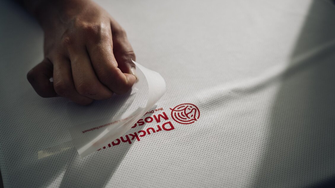 Eine Hand zieht die Transferfolie vom roten Druckhaus-Moser-Logo auf einem weißen Shirt ab. | © Bernd Grosseck