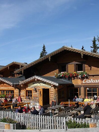 A cozy mountain hut with the sign "Eiskarhütte". Outside, guests are sitting on the terrace and enjoying the view of the surrounding mountains. | © Eiskarhütte
