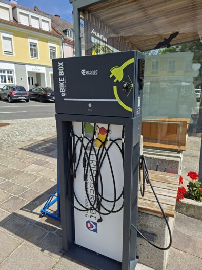 A modern bike box with charging sockets for e-bikes. It is located at a street corner, next to a bus stop.