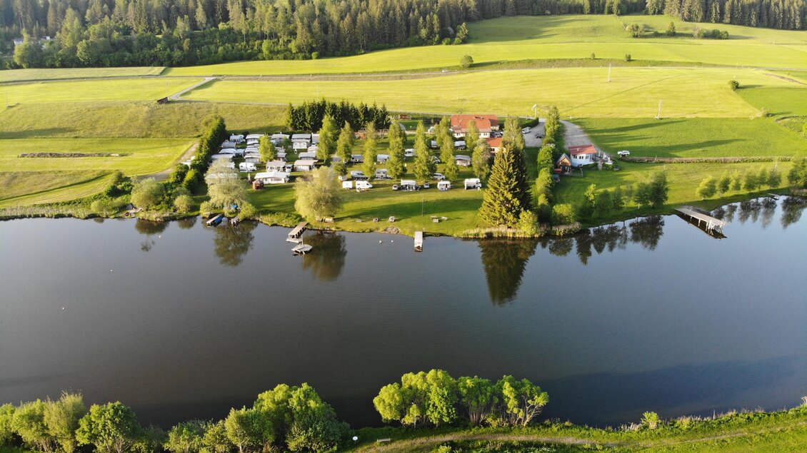 Eine ruhige Landschaft mit einem Fluss, umgeben von grünen Wiesen und Bäumen. Im Hintergrund sind Wohnmobile und ein kleines Gebäude sichtbar. | © Konrad Stückelberger