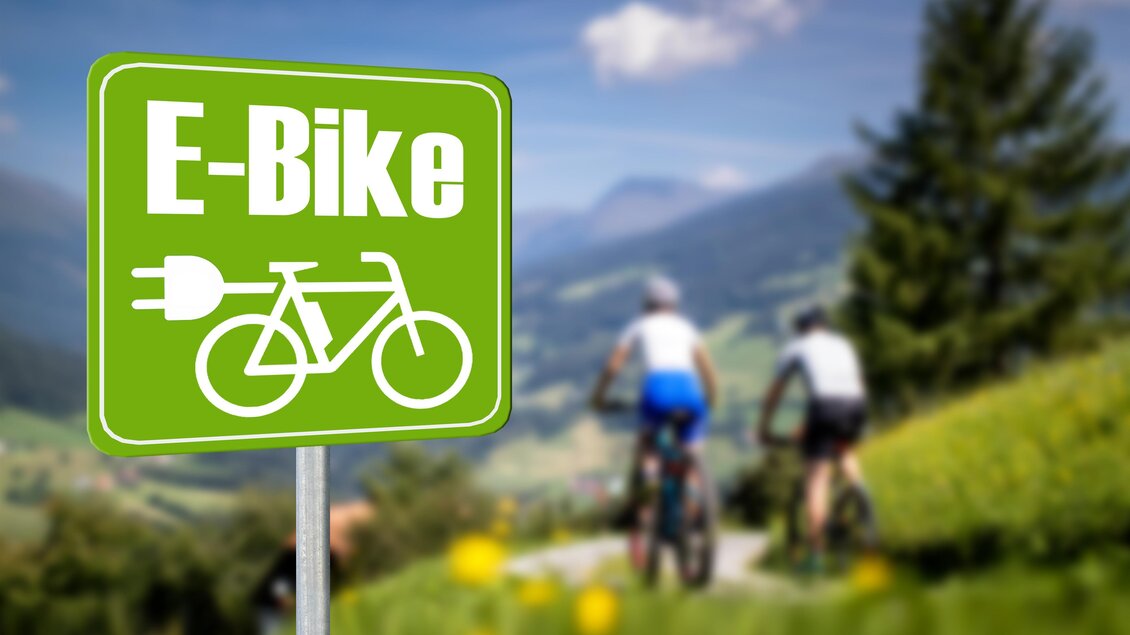 Ein Schild mit der Aufschrift "E-Bike" steht in einer grünen Landschaft. Im Hintergrund fahren zwei Radfahrer auf einem Weg. | © TV Murau