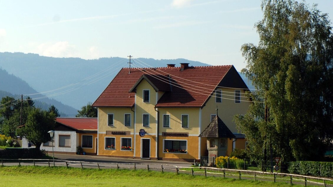 Ein hübsches gelbes Haus mit einem roten Dach und grünen Wiesen im Vordergrund. Im Hintergrund sind sanfte Hügel und ein blauer Himmel zu sehen. | © Roland Präsent