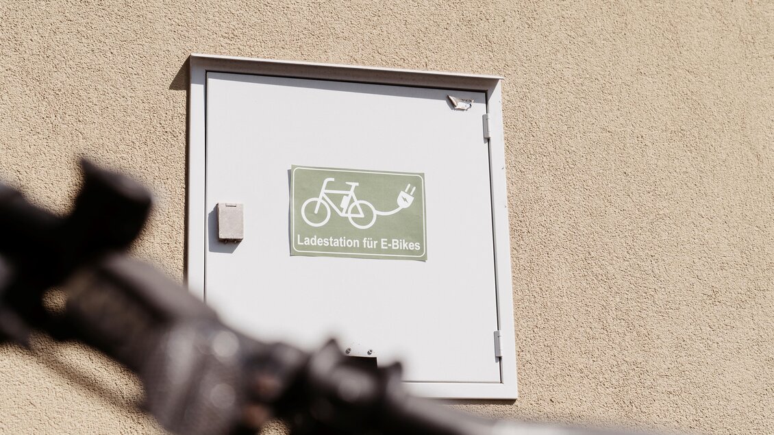 Eine Ladestation für E-Bikes ist an der Wand angebracht. Das Schild zeigt ein Fahrrad und ein Steckersymbol. | © Martinhof