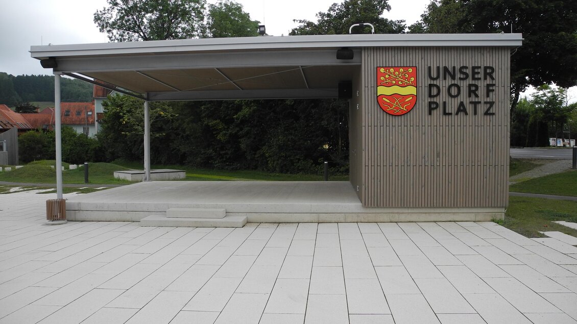 Ein moderner Pavillon mit dem Schriftzug "UNSER DORF PLATZ". Der Platz ist mit hellen Platten gestaltet und von Bäumen umgeben. | © Gemeinde Edelsbach bei Feldbach