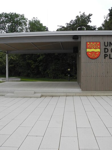 Ein moderner Pavillon mit dem Schriftzug "UNSER DORF PLATZ". Der Platz ist mit hellen Platten gestaltet und von Bäumen umgeben. | © Gemeinde Edelsbach bei Feldbach