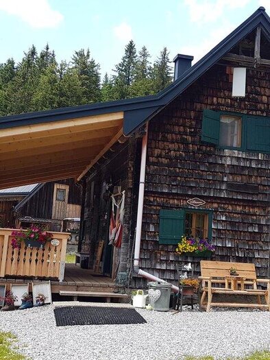 A cozy mountain cabin with wooden cladding and green shutters. The entrance is decorated with flowers and seating, surrounded by a forest. | © Scheikl Kordula