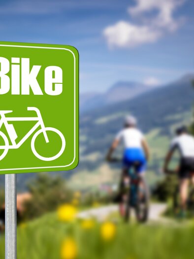 A green sign with the inscription "E-Bike" and a bicycle symbol. In the background, two cyclists can be seen on a beautiful hiking trail in a mountainous landscape. | © TVB Murau