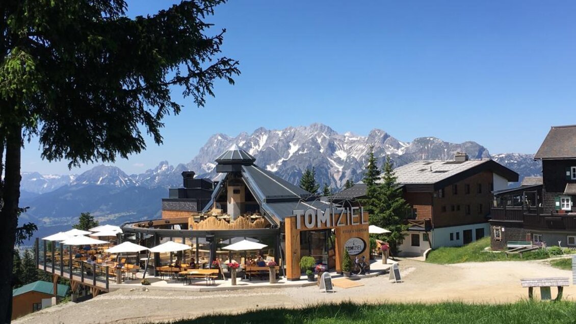 Ein alpin gestaltetes Restaurant mit einer Terrasse und Blick auf die Berge. Die Umgebung ist grün und sonnig, ideal für Erholung im Freien. | © TOMiziel