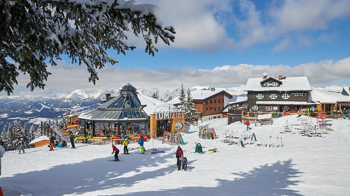 Ein schneebedecktes Bergdorf mit mehreren Gebäuden und Skifahrern. Der Himmel ist klar und die Landschaft ist malerisch.