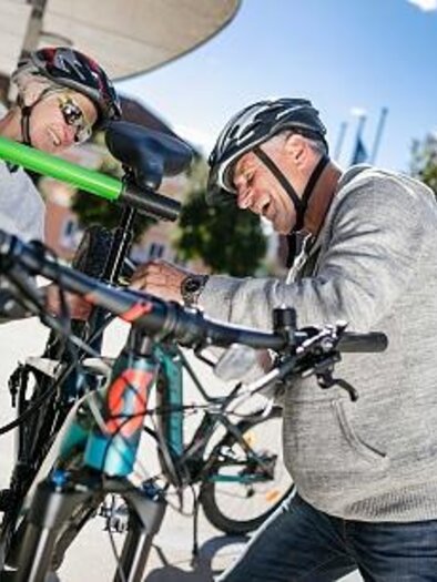 Zwei Personen reparieren zusammen Fahrräder an einer Wartungsstation im Freien. Die Sonne scheint und es sind Bäume im Hintergrund zu sehen. | © Thermen- & Vulkanland Steiermark, Harald Eisenberger