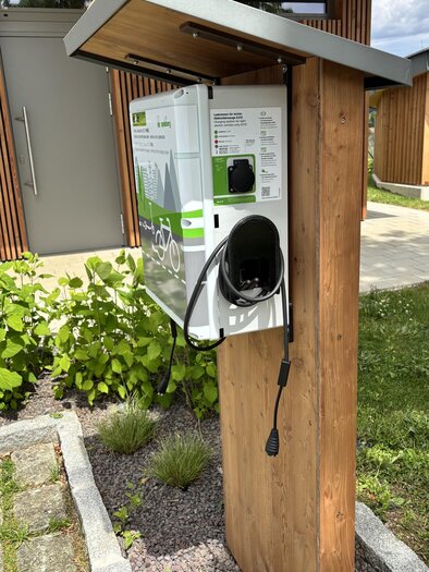 An electric vehicle charging station in a modern garden. It is mounted on a wooden post and surrounded by green plants. | © Marktgemeinde St. Nikolai im Sausal