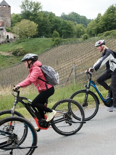Eine Gruppe von Radfahrern fährt auf einem Weg entlang von Weinbergen. Im Hintergrund ist eine Burg und grüne Bäume zu sehen. | © Schilcherland Steiermark
