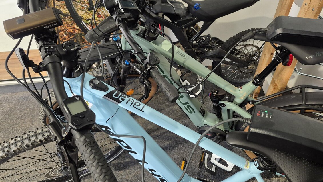 Zwei E-Bikes stehen nebeneinander in einem Raum. Die Fahrräder sind mit Kabeln verbunden und stehen auf einem Teppich. | © Andrea Stelzer