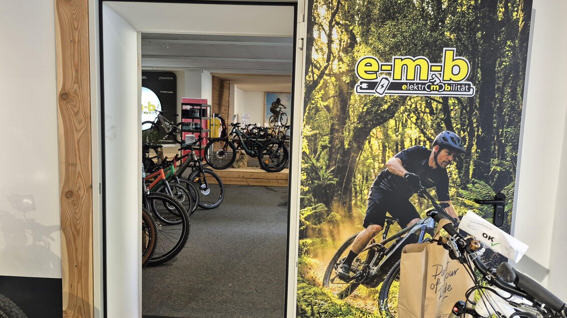 Ein Fahrradgeschäft mit einem einladenden Eingang. Im Hintergrund sind verschiedene Fahrräder und eine Wand mit einem lebendigen Motiv von einem Radfahrer in der Natur zu sehen. | © Andrea Stelzer