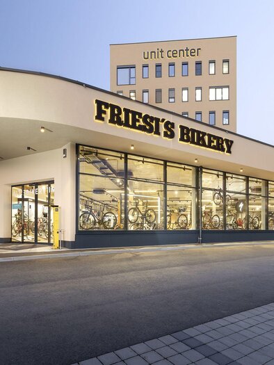 Eine moderne Bäckerei mit großen Fenstern und einem klaren Design. Im Hintergrund ist ein höheres Gebäude mit der Aufschrift „unit center“ zu sehen. | © Friesis Bikery
