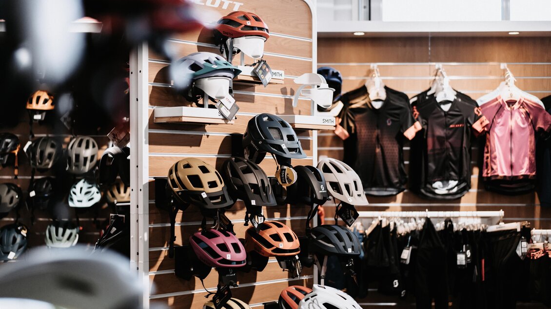 Ein Sportgeschäft mit einer Auswahl an Fahrradhelmen an einer Wand. Im Hintergrund hängen Fahrradbekleidung und Zubehör. | © Tourismusverband Oststeiermark