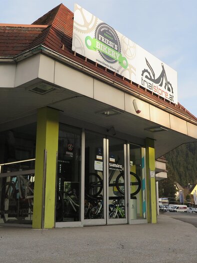 Ein Fahrradgeschäft mit großen Fenstern und bunten Plakaten. Im Hintergrund sieht man eine ruhige Straße und Bäume. | © Friesis Bikery