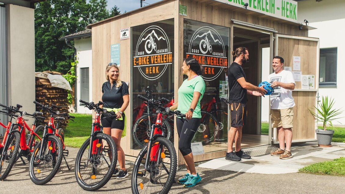 Ein Fahrradverleih mit mehreren Fahrrädern vor einer Holzlaube. Zwei Personen stehen an der Theke, während zwei weitere Personen auf der Seite warten. | © Herk Andreas