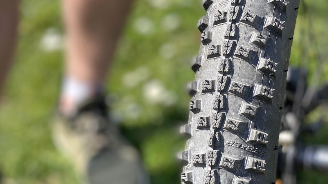 Ein naher Blick auf einen mountainbike Reifen mit grobem Profil. Im Hintergrund sind Beine einer Person in sportlicher Kleidung zu sehen. | © Ratscher Landhaus