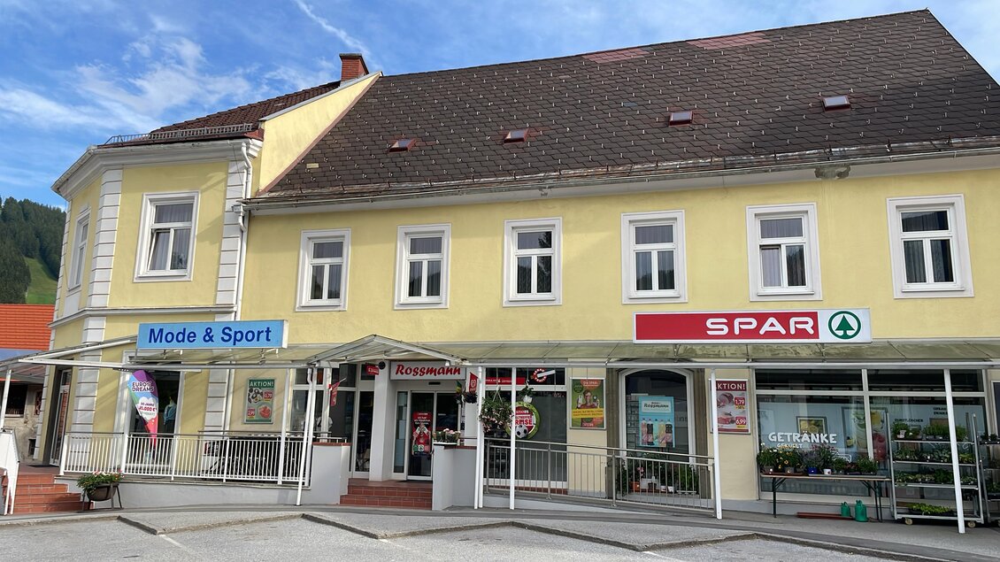 Ein gelbes Gebäude mit Geschäften, darunter ein Mode- und Sportgeschäft sowie ein SPAR-Supermarkt. Die Umgebung ist hell und freundlich, mit einem klaren Himmel. | © Doris Westphal