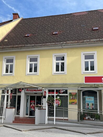 Ein gelbes Gebäude mit Geschäften, darunter ein Mode- und Sportgeschäft sowie ein SPAR-Supermarkt. Die Umgebung ist hell und freundlich, mit einem klaren Himmel. | © Doris Westphal
