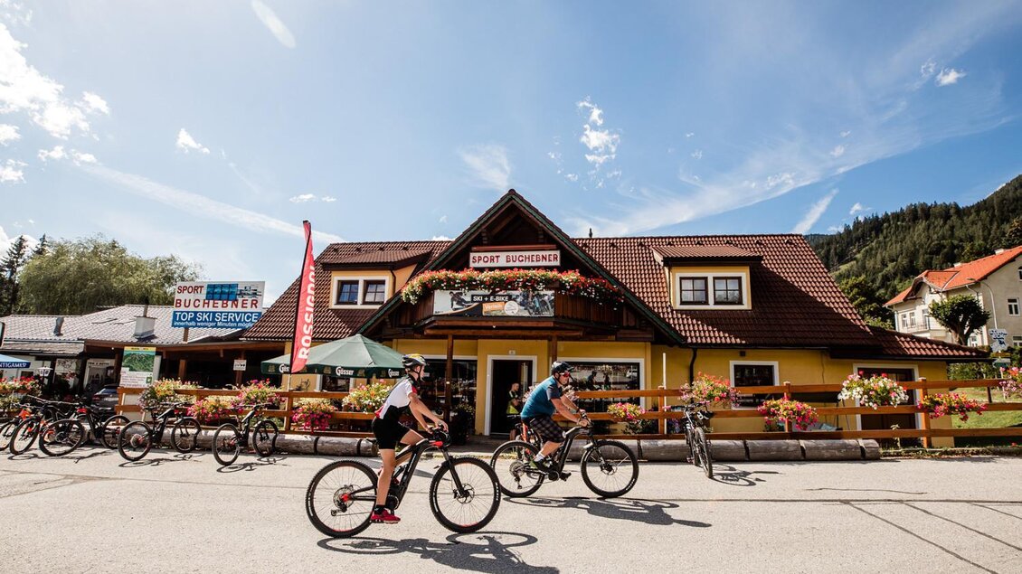 Ein malerisches Gasthaus mit einem roten Dach und bunten Blumen im Vordergrund. Zwei Fahrradfahrer fahren an der Straße vorbei, umgeben von einer schönen Landschaft.