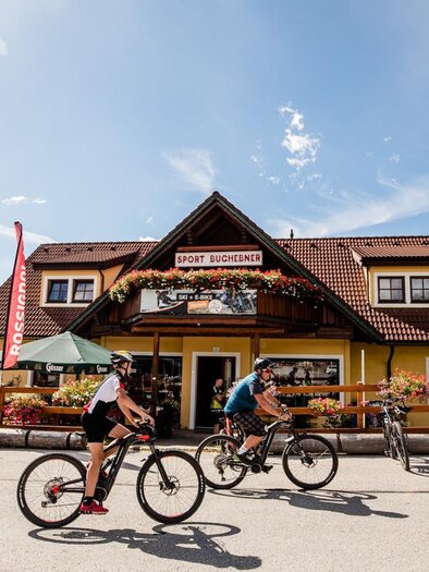 Ein malerisches Gasthaus mit einem roten Dach und bunten Blumen im Vordergrund. Zwei Fahrradfahrer fahren an der Straße vorbei, umgeben von einer schönen Landschaft.