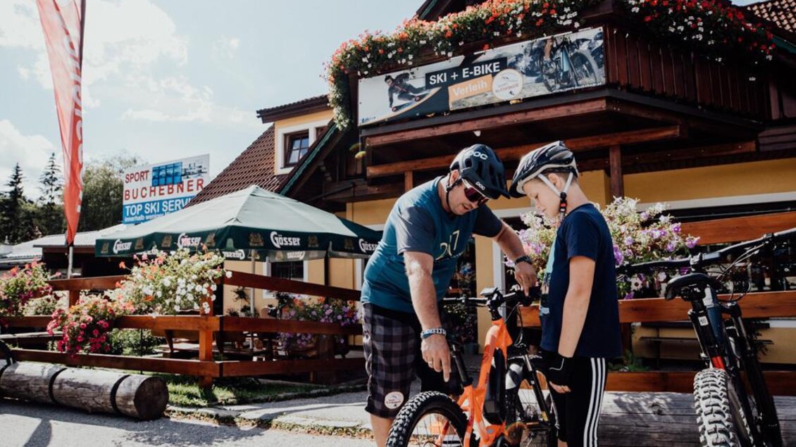 Ein Erwachsener und ein Kind stehen zusammen an einem Fahrrad. Im Hintergrund sieht man ein Gebäude mit Blumen und Sonnenschein.