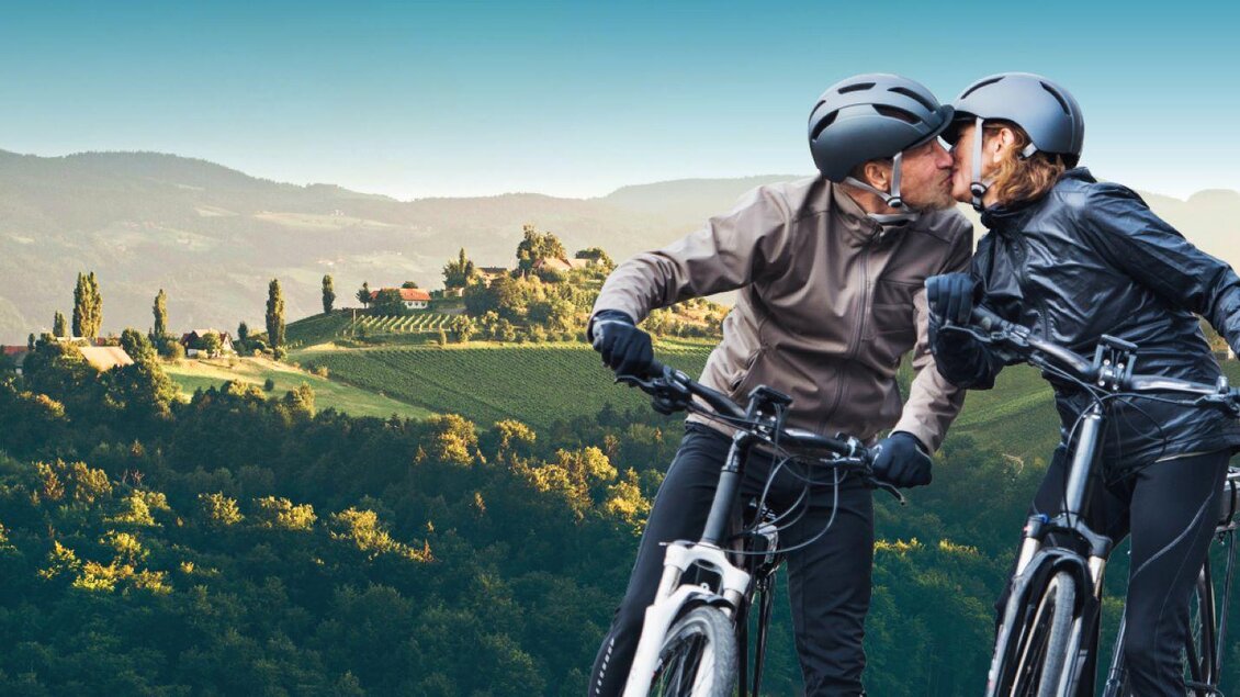 Ein Paar auf Fahrrädern küsst sich vor einer malerischen Landschaft mit Hügeln und Weinbergen. Die Szenerie strahlt Romantik und Abenteuerlust aus. | © E-Bike Visit Hotspot