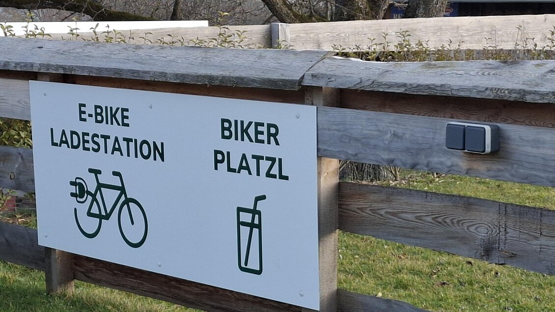 Eine E-Bike-Ladestation und ein Bereich für Radfahrer sind auf einem Holzschild angezeigt. Das Schild ist im Freien an einem Zaun befestigt.