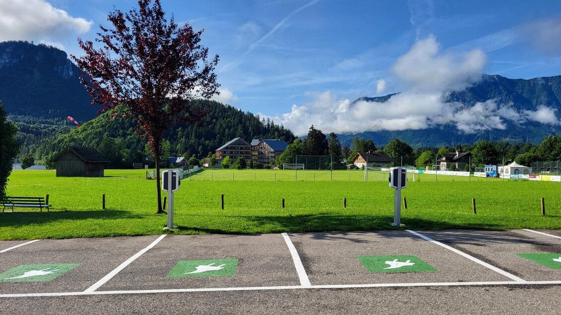 Eine weitläufige Wiese mit Bergen im Hintergrund. In der Vordergrund sind Parkplätze mit Ladestationen für Elektroautos zu sehen. | © Petra Kirchschlager
