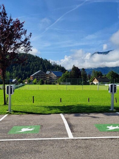 A spacious meadow with mountains in the background. In the foreground, there are parking spaces with charging stations for electric cars. | © Petra Kirchschlager