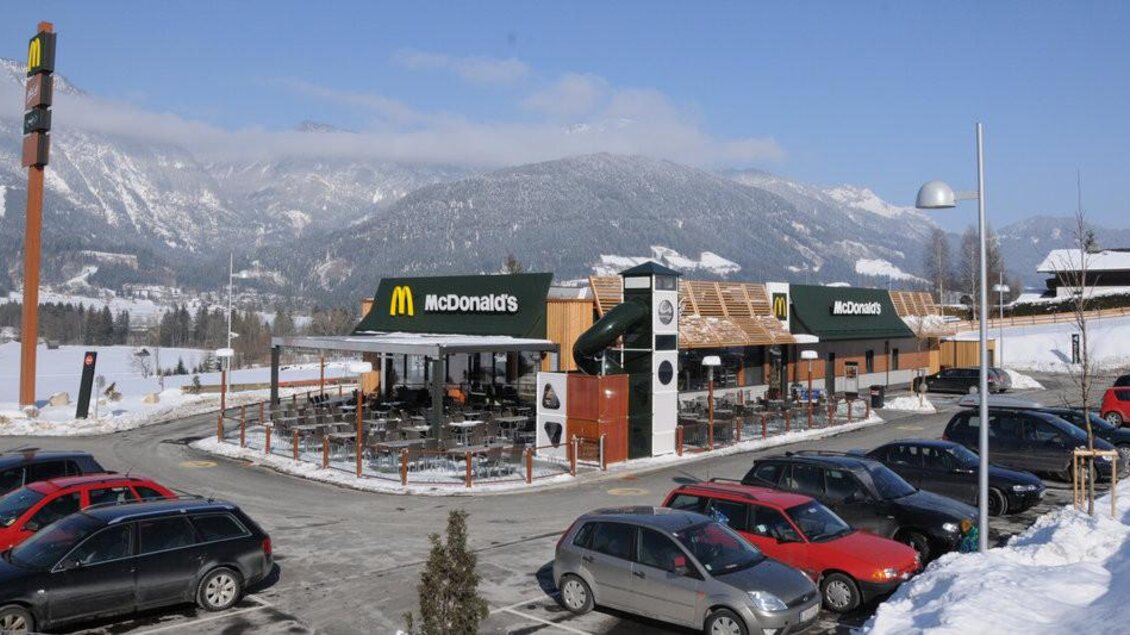 Ein McDonald's-Restaurant in einer schneebedeckten Landschaft mit Bergen im Hintergrund. Vor dem Restaurant steht ein Parkplatz mit mehreren Fahrzeugen. | © Hauser Kaibling