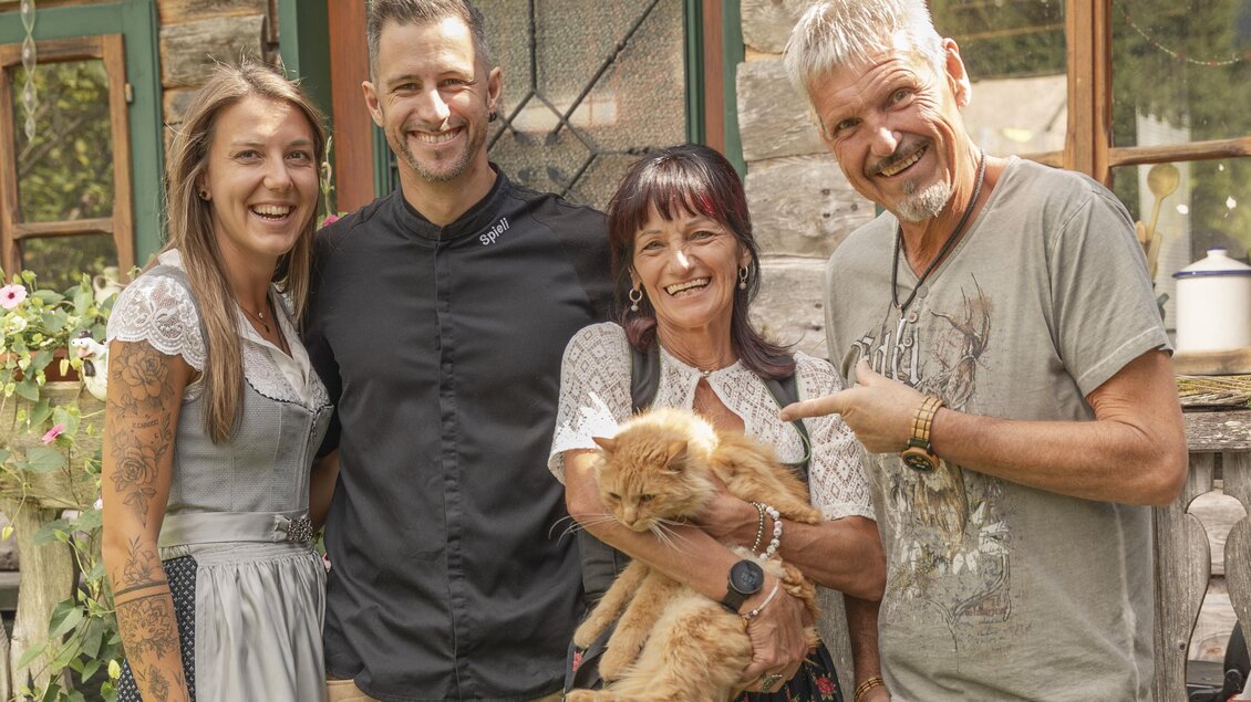 Eine Gruppe von vier Menschen posiert lächelnd vor einem Gebäude. Eine Frau hält eine Katze in ihren Armen und alle zeigen Freude und Zusammengehörigkeit.