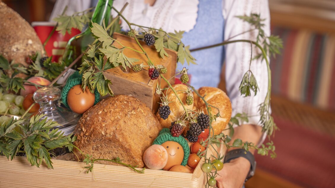 Ein Korb voller frischer Lebensmittel, darunter Brot, Eier und Beeren. Das Arrangement ist mit grünen Pflanzen dekoriert.