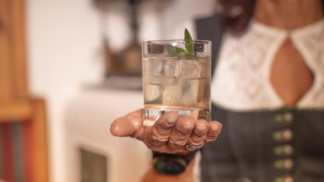 Ein Glas mit einem klaren Getränk und Eiswürfeln wird von einer Person gehalten. Im Glas schwimmt ein grünes Minzblatt.
