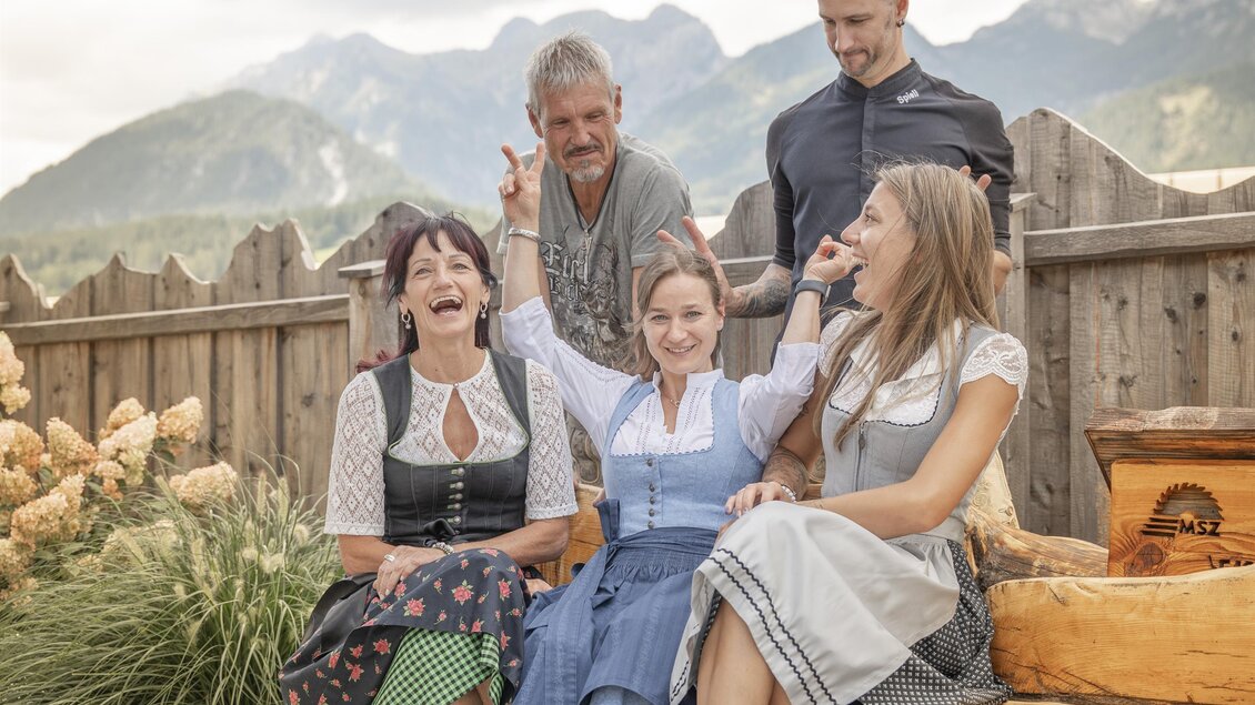 Eine Gruppe von fünf Personen posiert fröhlich im Freien, umgeben von einer naturbelassenen Landschaft. Im Hintergrund sind Berge und eine Holzprivatsphäre zu sehen.
