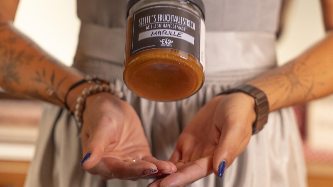Eine Person in traditioneller Tracht hält ein schwebendes Glas mit einer goldenen Flüssigkeit. Der Hintergrund ist unscharf und betont das Glas im Vordergrund.