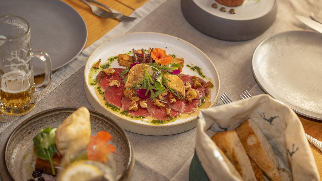 Ein elegantes Essen auf einem Tisch, mit zartem Carpaccio, frischen Kräutern und bunten Garnituren. Am Tisch stehen außerdem verschiedene Vorspeisen und ein Bierglas.