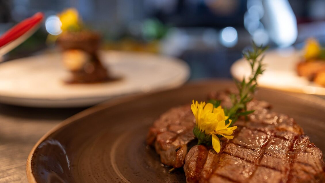 Ein saftiges Steak auf einem braunen Teller, garniert mit einer Blume und frischen Kräutern. Im Hintergrund sind weitere Gerichte sichtbar.