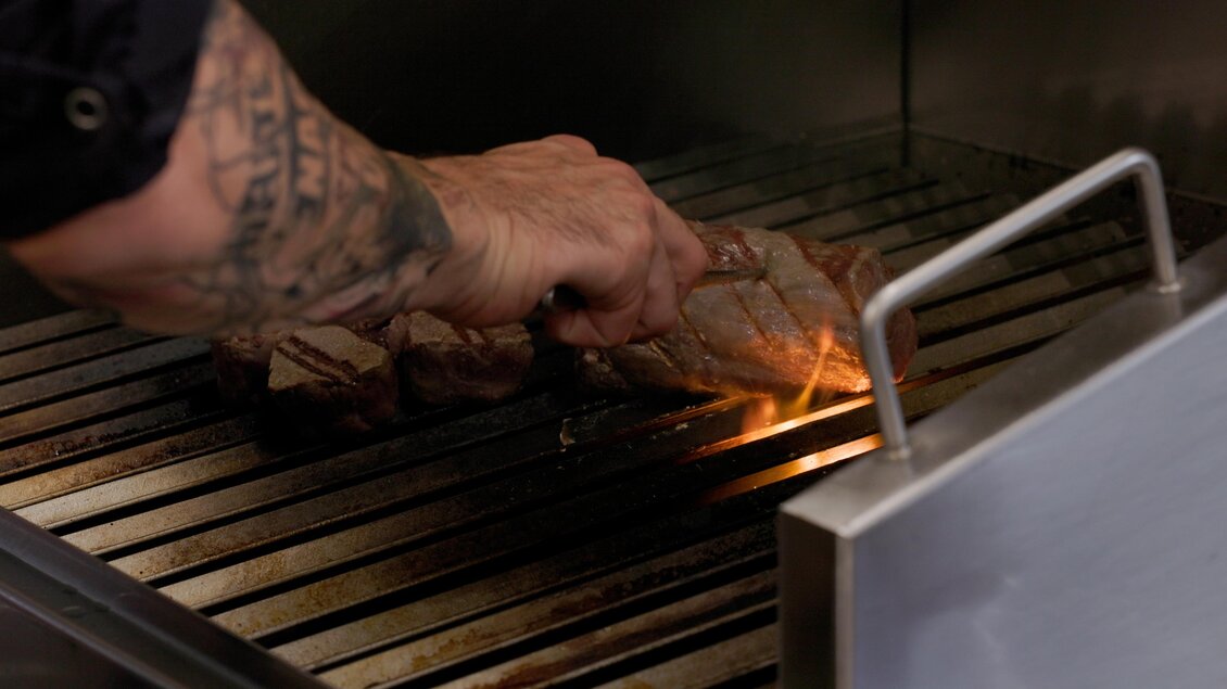 Ein Koch grillt ein Stück Fleisch auf einem Grill. Flammen züngeln auf, während er das Fleisch wendet.