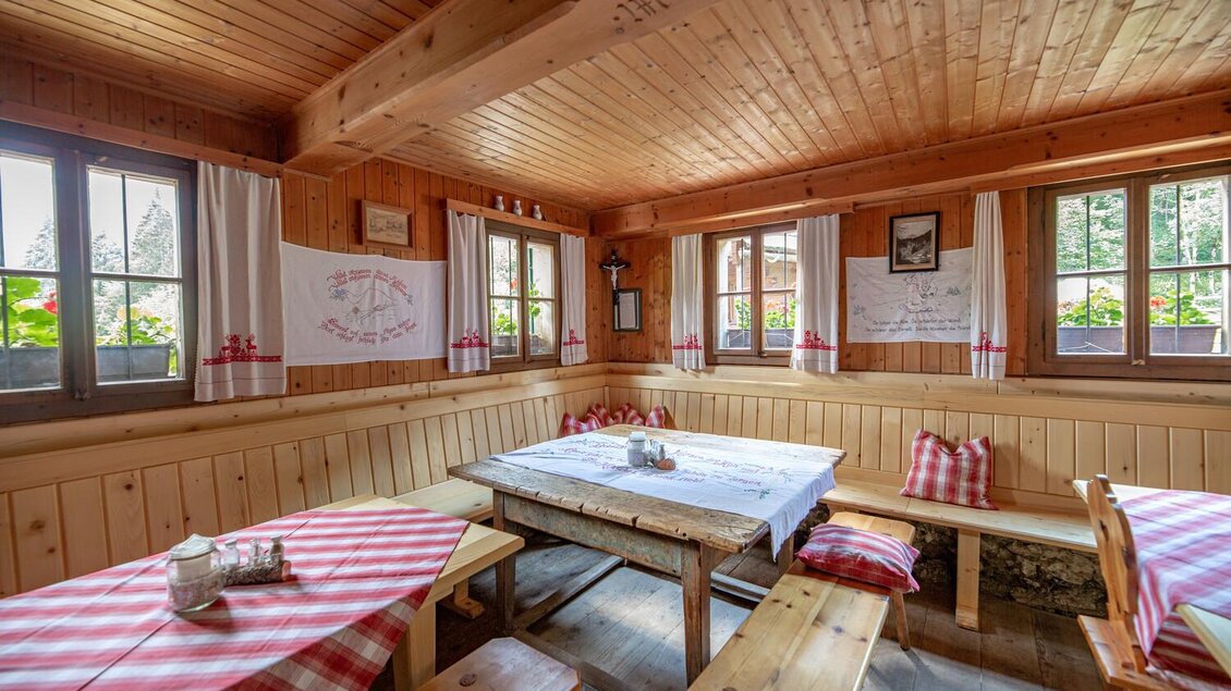 Eine gemütliche Stube mit Holzverkleidung und rustikalen Bänken. Auf den Tischen liegen rot-weiß karierte Tischtücher und die Fenster sind lichtdurchflutet. | © TV Gesäuse
