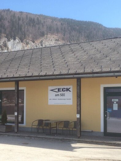 A building with a yellow facade and a flat roof. In front of the entrance, there are tables and chairs, and in the background, you can see mountains. | © Waltraud Loitzl