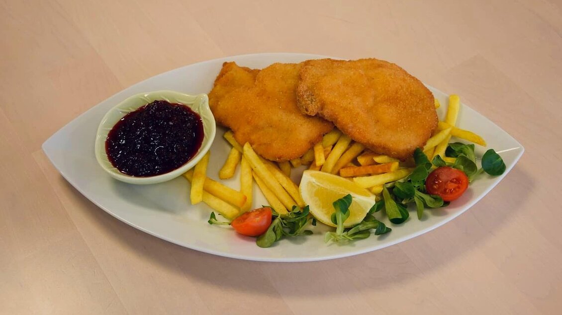 Ein Teller mit zwei panierten Schnitzeln, Pommes frites und frischen Beilagen. Dazu gibt es ein Schälchen mit Preiselbeeren. | © Edenhof