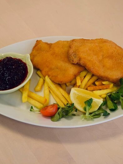 Ein Teller mit zwei panierten Schnitzeln, Pommes frites und frischen Beilagen. Dazu gibt es ein Schälchen mit Preiselbeeren. | © Edenhof